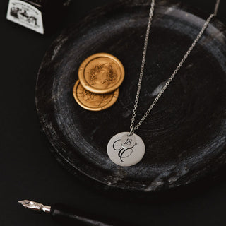 Silver personalised disc necklace with engraved script initial and small “18” charm on a chain, styled on a dark decorative tray with wax seals and a fountain pen nib.