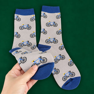 A pair of crew socks with a repeating bicycle pattern on a light gray background, featuring blue cuffs, heels, and toes, held by a hand against a solid dark green background.