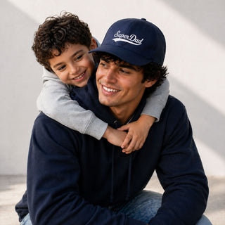 Smiling father wearing a navy Super Dad cap with child hugging him from behind, celebrating Father’s Day in a bright studio setting