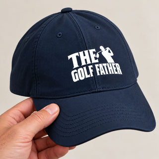 A navy blue baseball cap with a white embroidered graphic reading 'THE GOLF FATHER' and a golfer silhouette, held by a hand against a plain light beige background.