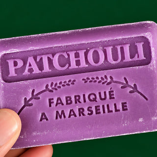 A close-up of a purple patchouli bar soap held by a hand against a solid dark green background. The soap is embossed with the brand name 'PATCHOULI' and 'FABRIQUÉ A MARSEILLE'.