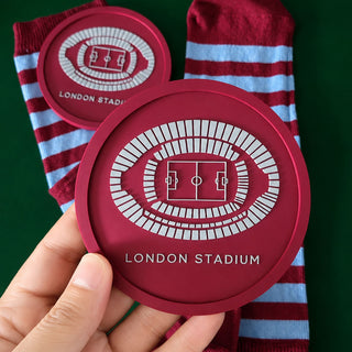 A hand holds a round red coaster featuring a white stadium floor plan and the text 'LONDON STADIUM', with two striped socks in the background against a dark green backdrop.