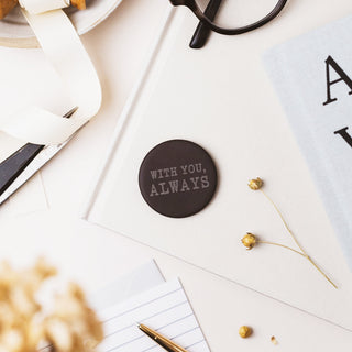 A round dark brown coaster with the words 'WITH YOU, ALWAYS' printed in a light gray serif font, styled in a flat-lay arrangement with glasses, a notebook, dried flowers, and a ribbon.