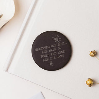 A round black coaster with silver engraved text reading 'WHATEVER OUR SOULS ARE MADE OF YOURS AND MINE ARE THE SAME' and small starburst motifs, placed on a white notebook with dried flowers nearby.