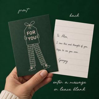 A hand holds a dark green greeting card and a lined note card against a dark green background. The card has a simple line drawing of a person holding a gift tag that says 'FOR YOU.