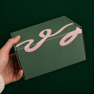 A hand holding a dark green cardboard box with a large pink abstract script logo on the top, set against a solid dark green background.