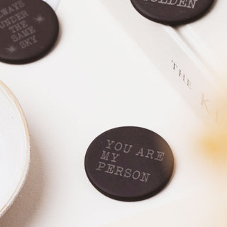 A close-up of a round black coaster with the phrase 'YOU ARE MY PERSON' in light gray lettering, placed on a white surface next to other similar coasters.