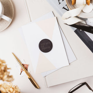 A styled flat-lay of a white envelope with a geometric beige design and a round dark brown sticker that reads 'YOU ARE MY PERSON'. The envelope is surrounded by stationery props including a gold pen, a notebook, a coffee cup, and dried flowers on a light beige surface.
